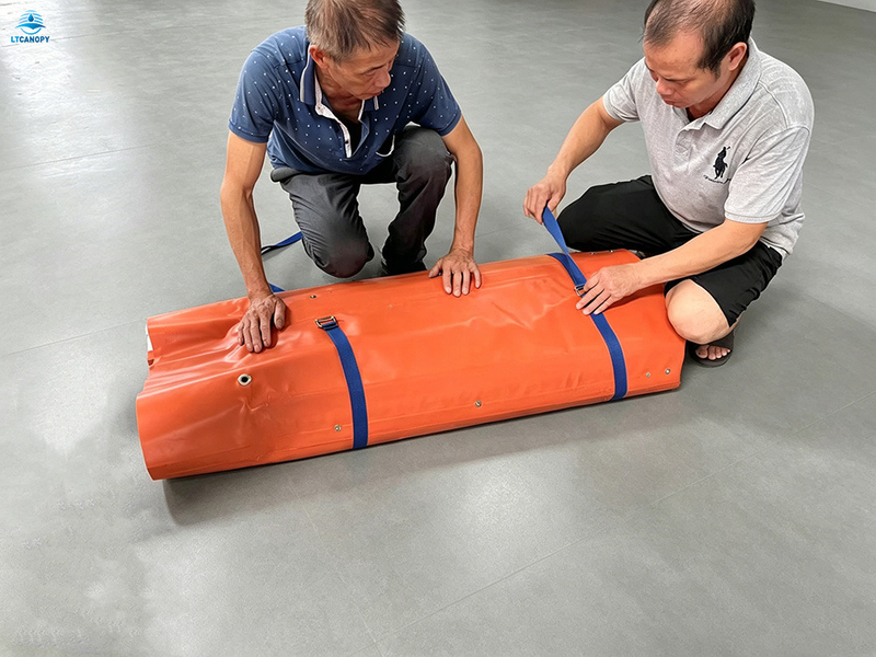 Workers securing rolled-up orange flood barrier with straps for compact storage and transport